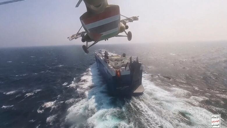 The view of a helicopter, from below, as it flies toward a merchant vessel on the sea.