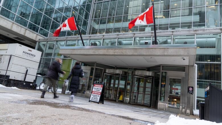 Two people walk into a highrise office building in winter. There are big Canada flags over the entrance.