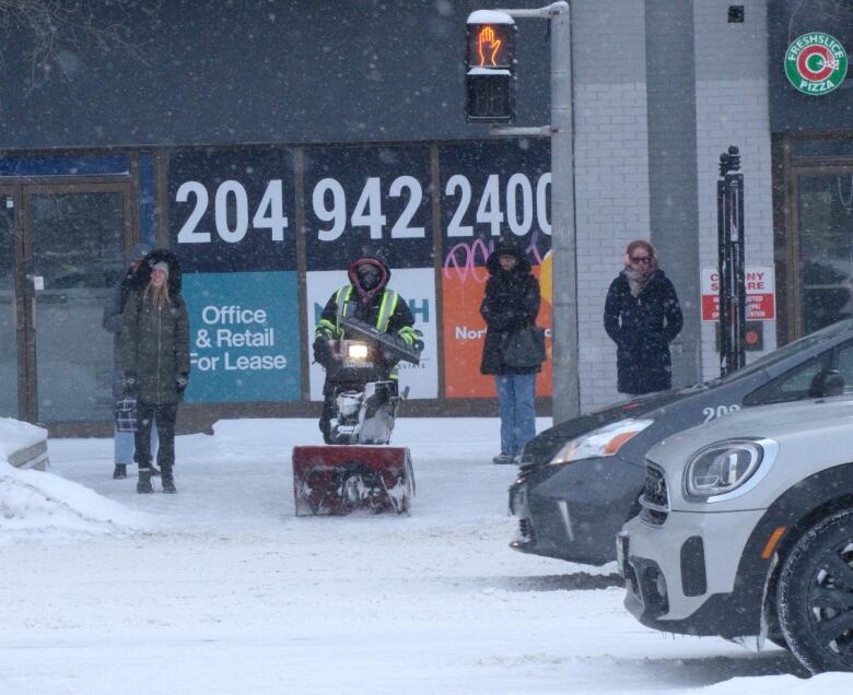 People, including someone pushing a snowblower, prepare to cross a street at a crosswalk.