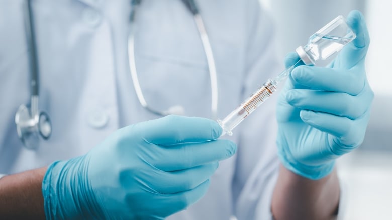 A photo of a medical worker with a stethoscope holding up a syringe going into a bottle.