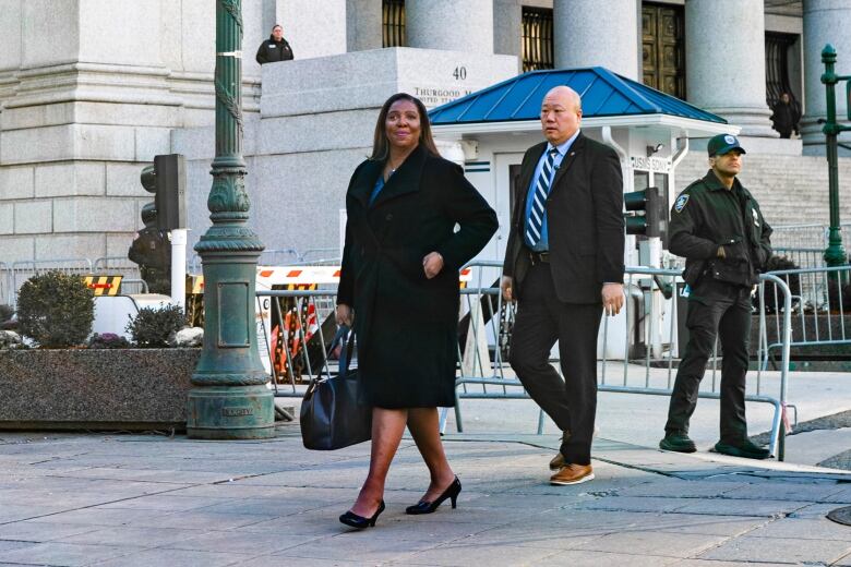 A woman in a long black coat and a man in a black suit walk past a large, concrete building.