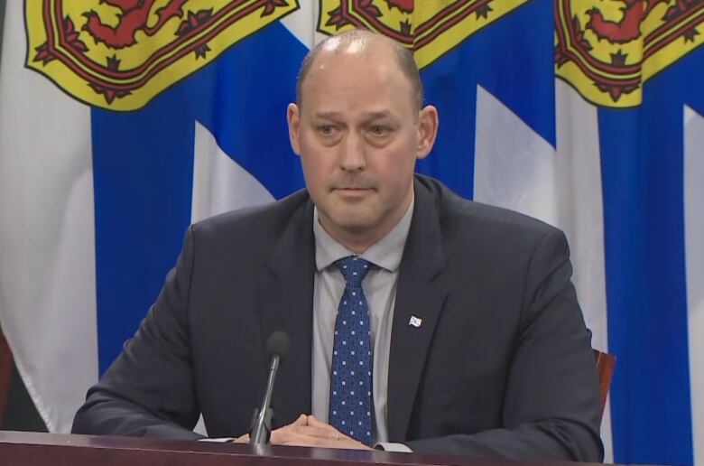 A bald man sits at a table with his hands folded against the backdrop of Nova Scotia flags.