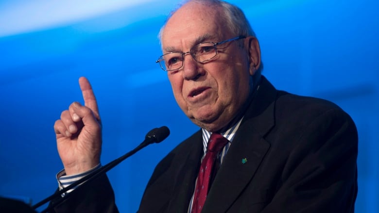 A man wearing glasses stands in front of a microphone with one of his hands in the air. 