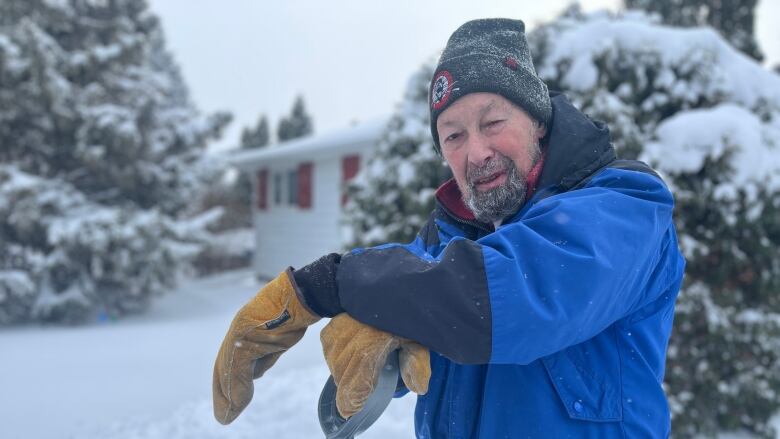 A man in a winter jacket, toque and large mitts leans over a snow shovel.