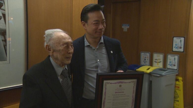 Two men stand next to each other smiling, one holding a declaration 