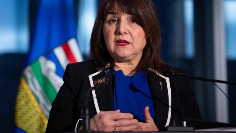 A woman with a black suit and blue blouse stands at a podium. 