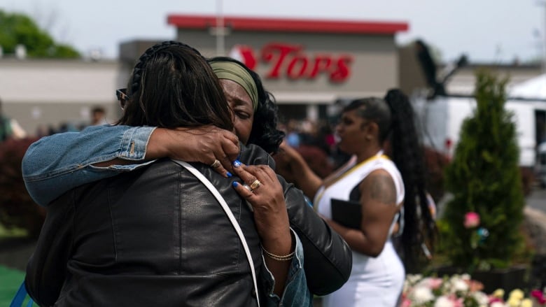 Two women embrace outside, with a building in the background that says, 'Tops.'