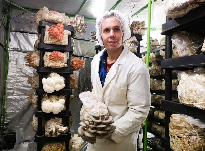 A mushroom grower holds a blue oyster mushroom while standing in front of racks of other species of fungi.