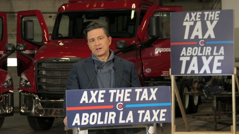 A man in a dark blazer and blue turtleneck sweater stand behind a podium and in front of two red semi-truck trailer cabs.