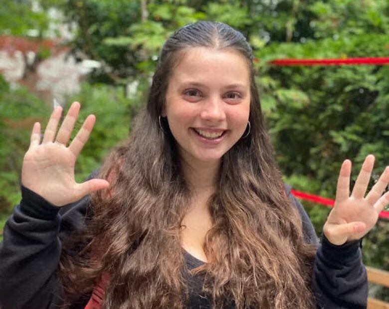 A girl with long brown hair and wearing a black hoodie is smiling and holding up nine fingers. 