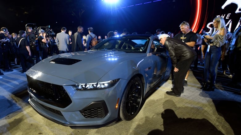 A crowd of people look at a blue-grey car.