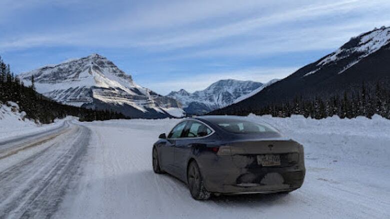 A blue tesla is on the side of a snow covered road. In the distance is a large mountain blanketed in snow. 