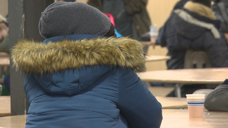 The back of a person sitting at table in a blue parka with a fur-lined hood is seen.