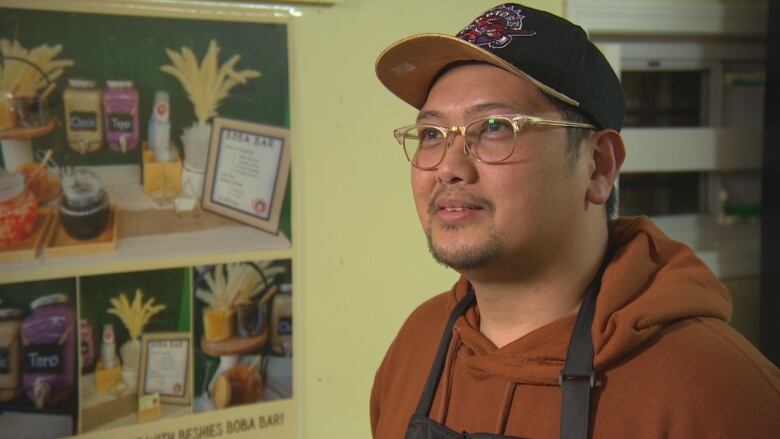 A man in a orange sweater, black apron and ball cap is standing as he is being interviewed.