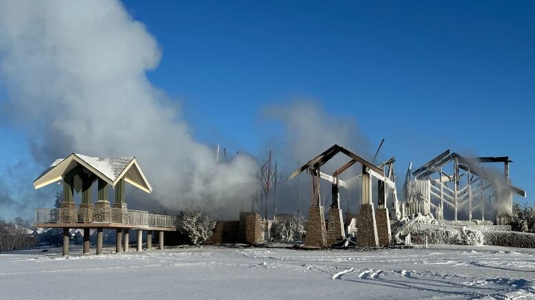 The burned remnants of a building stand in fresh snow, under a clear blue sky, as smoke floats from the scene.