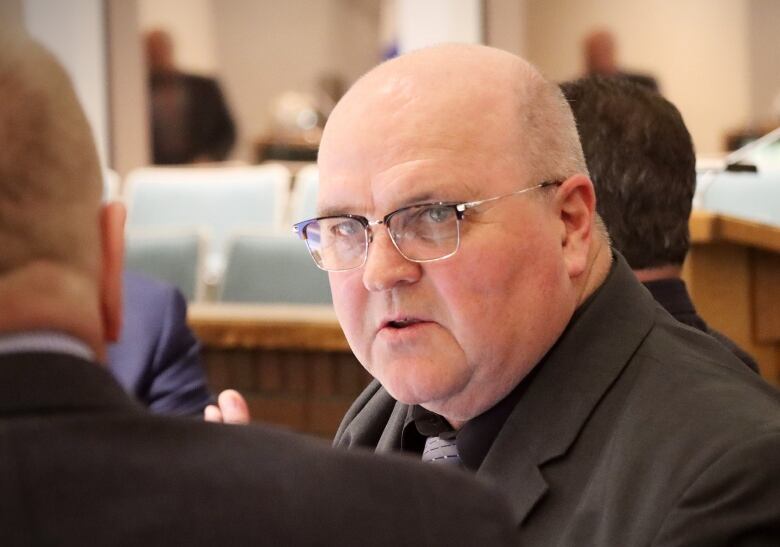 A balding man with glasses wearing a dark suit talks with someone whose back is to the camera.