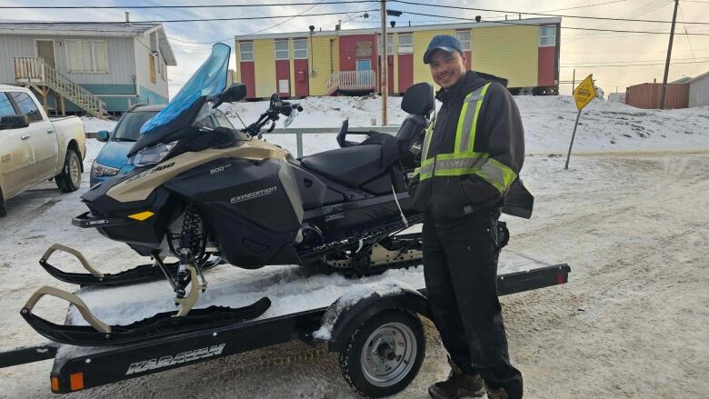 A man in black clothing with reflective yellow safety stripes stands in front of a small flatbed trailer with a snowmobile on it.