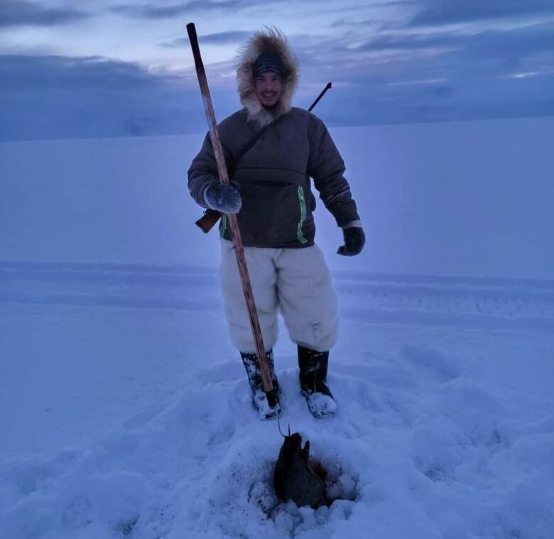 A man, standing on vast ice, in what appear to be fur pants and a fur lined hooded parka holding a harpoon up from a hole in the ice. The harpoon appears to be holding up a seal from that hole.