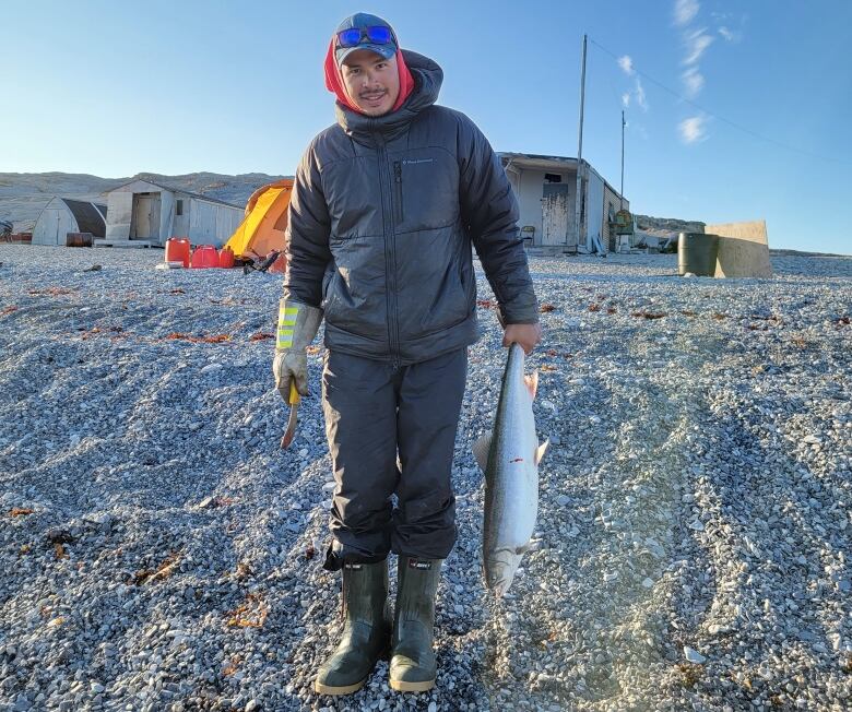 A man stands holding a fish on vast rocky land. A few small structures can be seen behind him. He's wearing black clothing, a ball cap, and sunglasses, and a knife in his right hand.