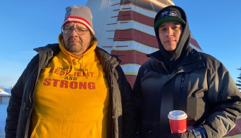 Two men stand outside in the cold in front of a teepee. 