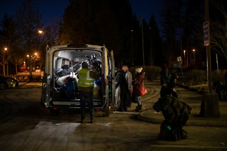 A man stands with his back to the camera wearing a safety reflector vest and black pants. It is dark outside and he is on a street facing into the open back doors of a white van and appears to be packing something into the vehicle. Around the van is a handful of people, one of whom appears to be carrying garbage bags of personal belongings.