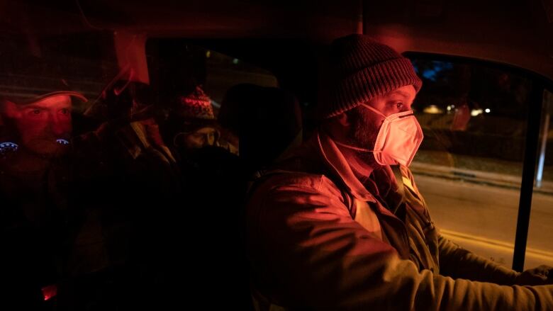 A man wears a medical mask and a toque while sitting in the driver's seat of a vehicle. It is dark outside and there is the silhouette of people sitting behind him in the vehicle.