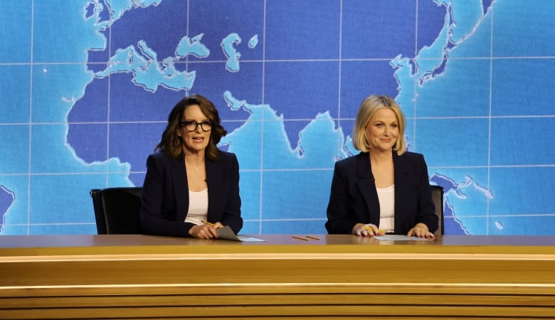 From left, a woman with brown hair and a woman with blonde hair are shown seated at a desk.