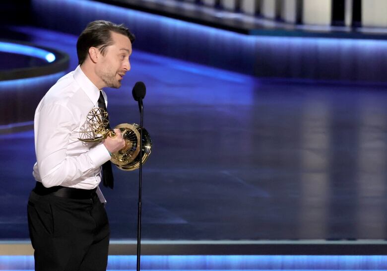 A man holds a golden statuette and speaks into a microphone, looking off-stage during an awards show.