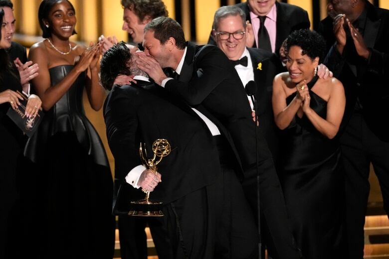 Two men in tuxedos kiss as one holds an Emmy award while people around them clap and smile. 