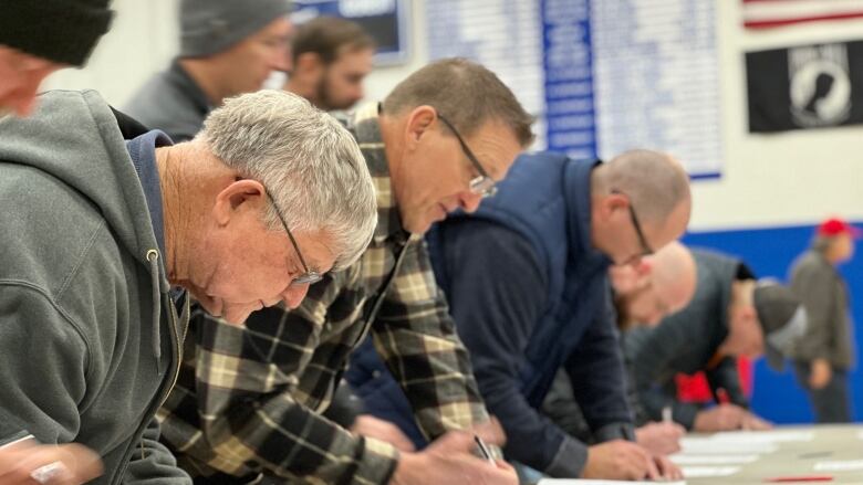 Men line lean over a table, writing on pieces of paper on a table.