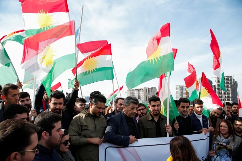 Several people hold up a banner while others hold flags at an outdoor demonstration.