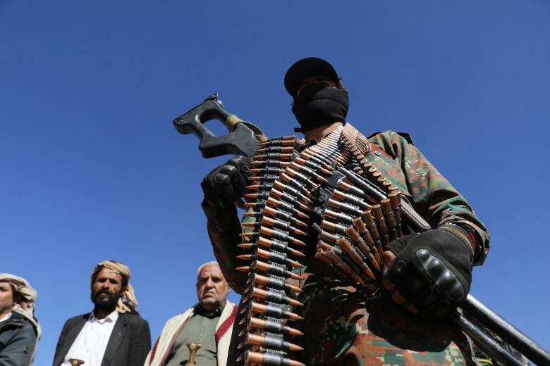 A man with his face covered stands hold a machine gun.