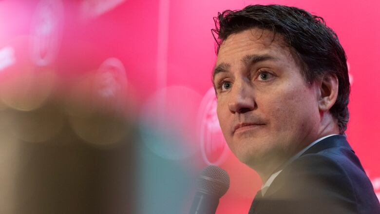 A man in a suit looks into a crowd while holding a microphone. He is sitting in front of a red backdrop.