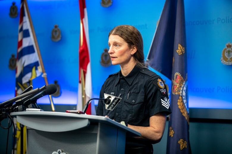 A woman in police uniform is pictured at a podium with mics, behind her the B.C., Canada, and police flags.