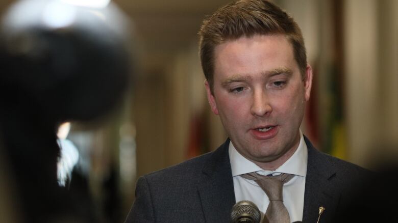 A man in a dark grey suit, white shirt and brown tie speaks into a microphone. 