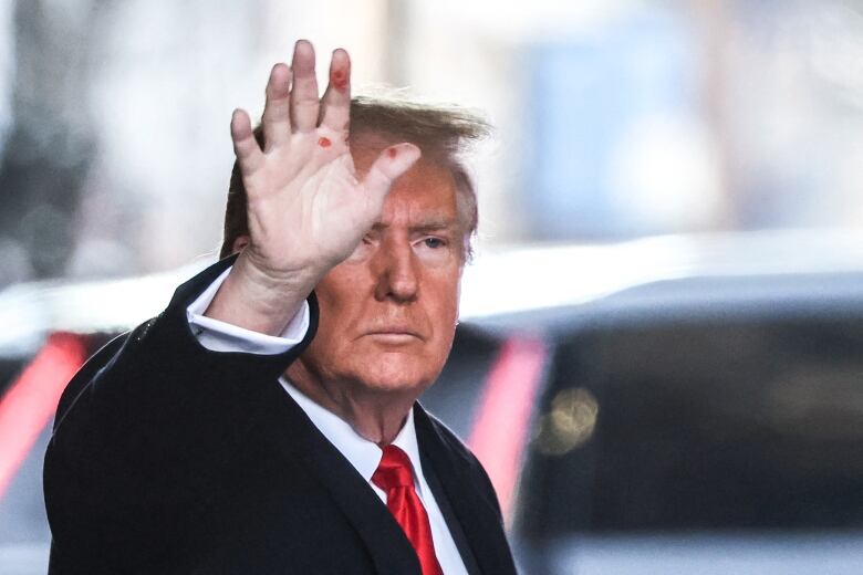 An older, cleanshaven man wearing a tie and a dark winter coat waves to unspecified persons off camera. He appears to have cuts or abrasions on his right hand.