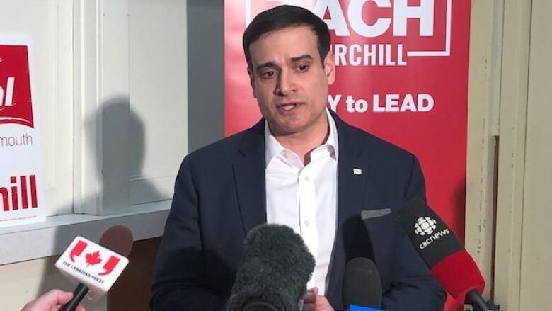 A man in a black suit and white shirt speaks to reporters.