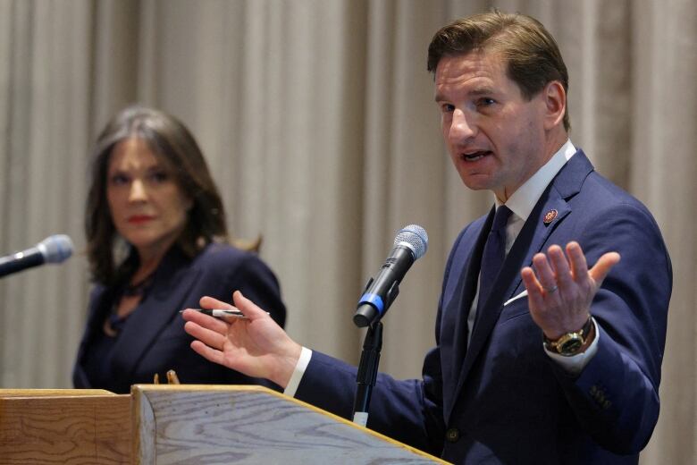 A clean-shaven man in a suit and tie speaks at a podium as a woman in a suit blazer looks on.