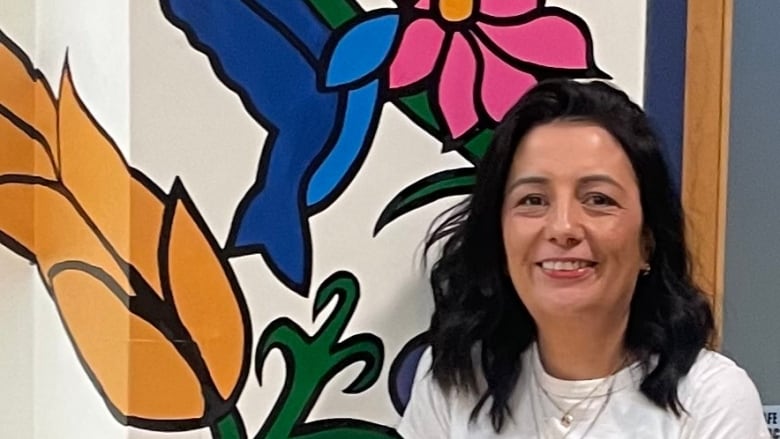 A woman stands and smiles at the camera. She's in a school setting, standing in front of flowery wallpaper.