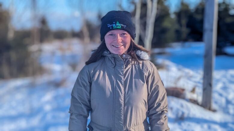 A woman wearing a toque and winter jacket stands in a snowy treed area. 