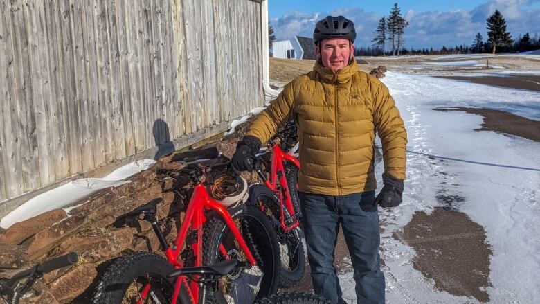 A man wearing a yellow winter jacket and helmet stands next to two red fat bikes. 