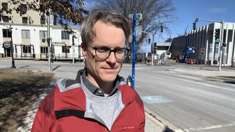 Man with greying hair and black glasses stands on city sidewalk wearing casual red jacket.