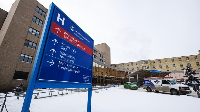 A blue sign with arrows and directions showing how to access a hospital is shown in the foreground with a brick hospital building in the background surrounded by a snow-covered ground, on an overcast winter day.