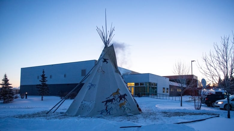 A teepee with smoke emnating from it sits in front of a building at dawn on a cold winter day.