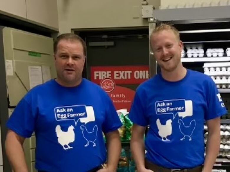 Two men in matching blue t-shirts stand in front of a cooler full of eggs.