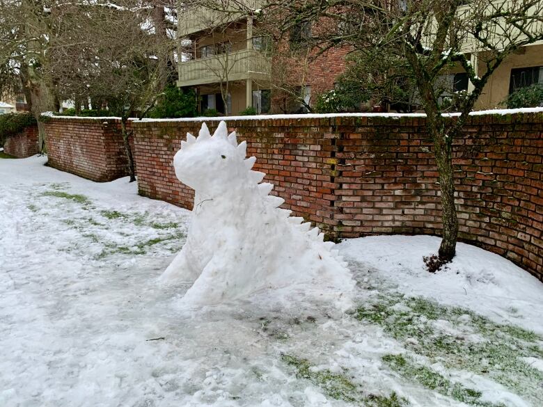 A dinosaur-shaped snow sculpture next to a brick wall.