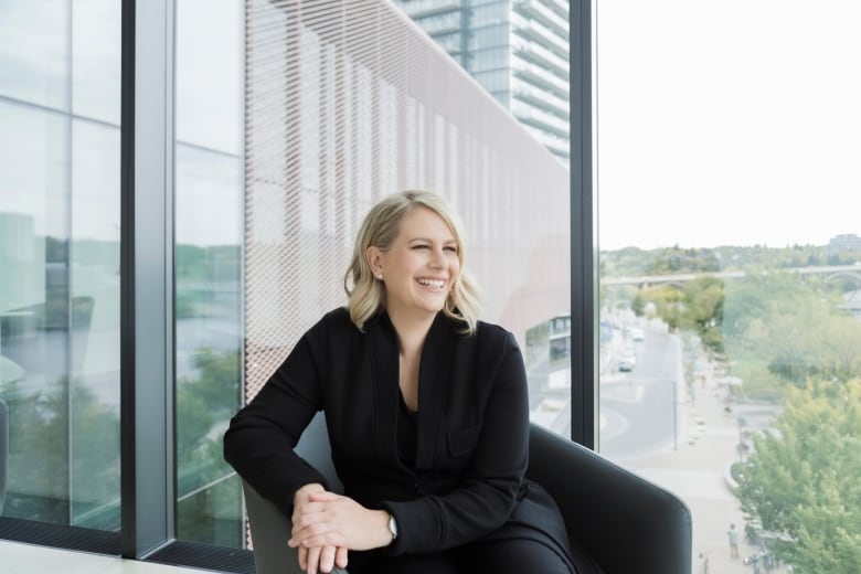 A woman in a black blazer looks off camera as she smiles. She has blonde hair and is sitting on a black chair. She's sitting in front of a window