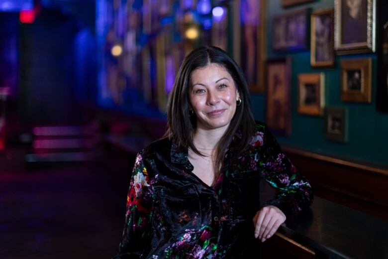 A brunette woman wearing a black button down with floral print leans against a dark wooden bar that's attached to a wall. The wall is painted dark green and is filled with framed photographs.