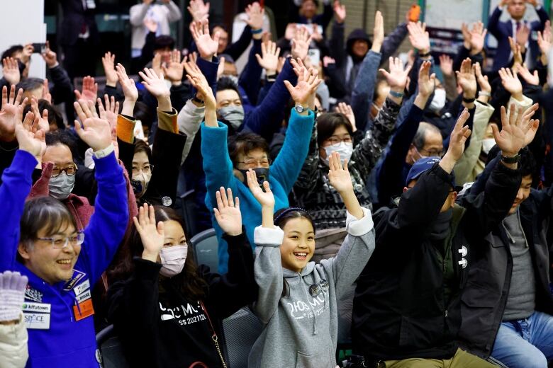 Dozens of people, both adults and children, are shown lifting their arms in a public setting. Some are wearing COVID masks.