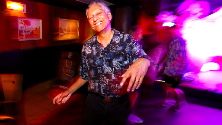 A smiling man dances in a colourfully lit room. The background is blurry. 